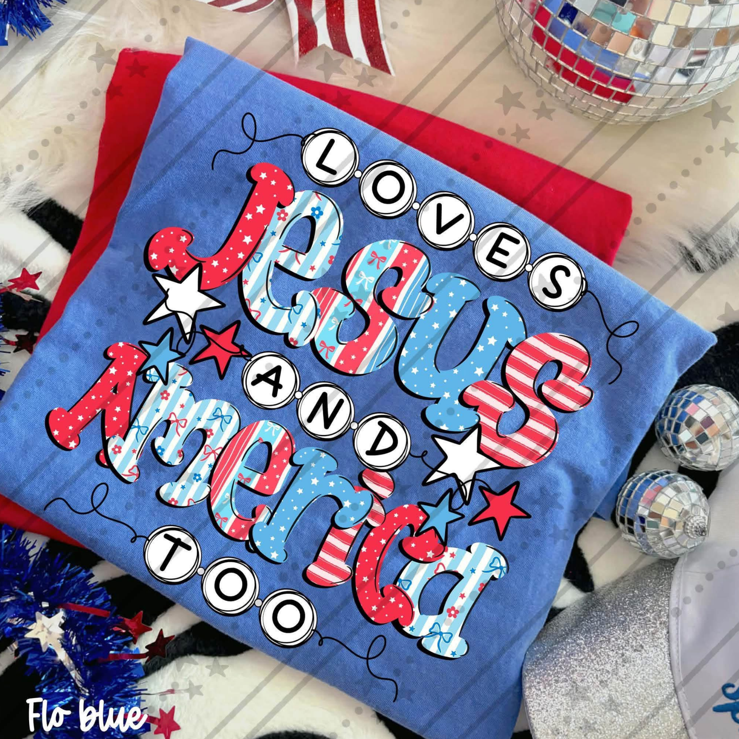 Blue Comfort Colors® graphic t-shirt featuring the phrase "Loves Jesus and America Too" in colorful patriotic lettering filled with stars, stripes, and bow patterns.