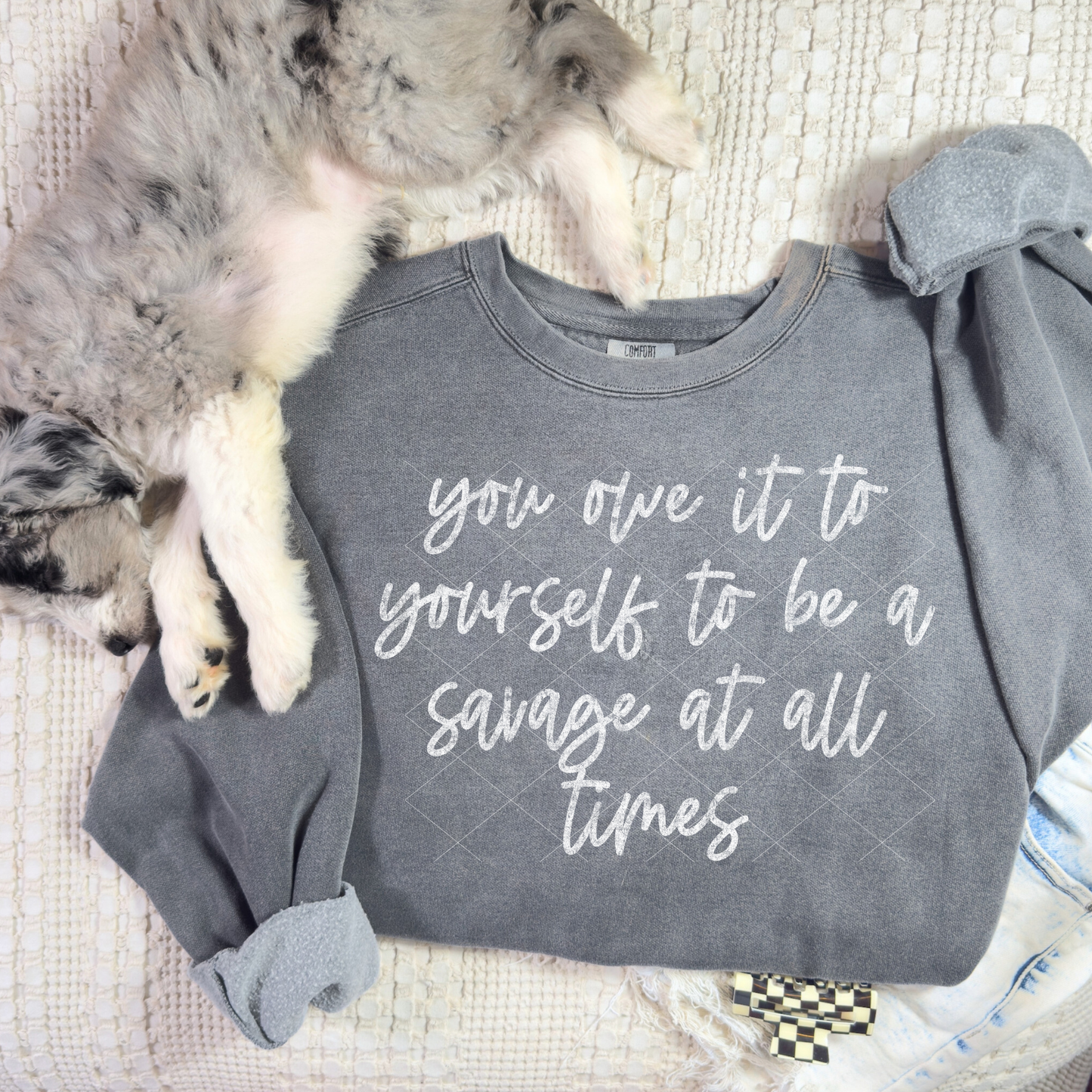 Gray sweatshirt with the phrase ‘you owe it to yourself to be a savage at all times’ displayed flat with a dog laying beside it.