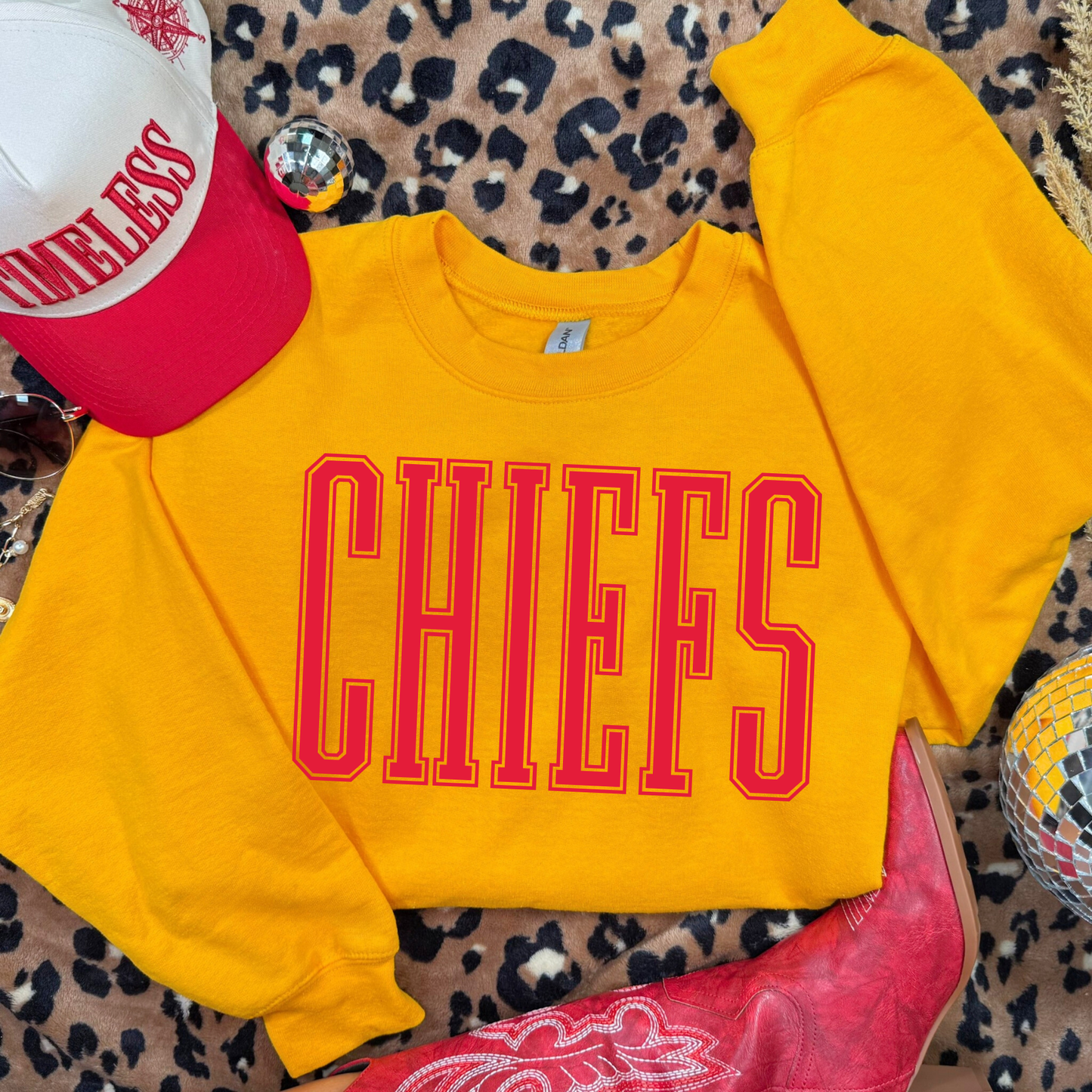 Yellow sweatshirt with 'CHIEFS' text, red cowboy boots, and a red and white cap on a leopard print surface.