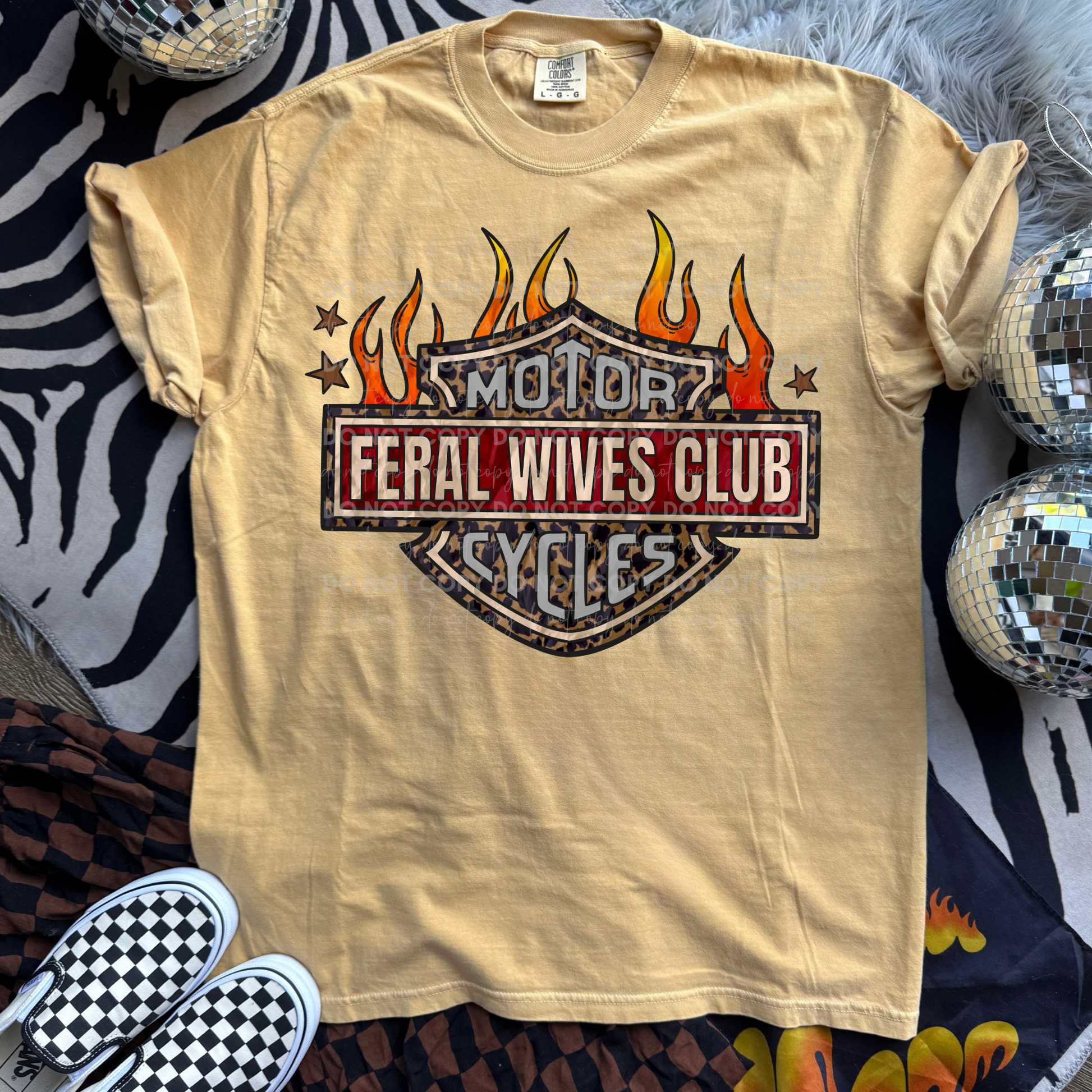 Feral Wives Club graphic tee featuring a vintage motorcycle club-inspired emblem with leopard print detailing, bold red lettering, orange flames, and distressed biker-style artwork printed on Comfort Colors shirts.