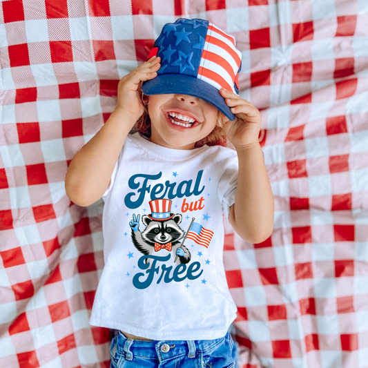 Child wearing a white Feral but Free patriotic raccoon graphic t-shirt with American flag design and red, white and blue holiday theme.