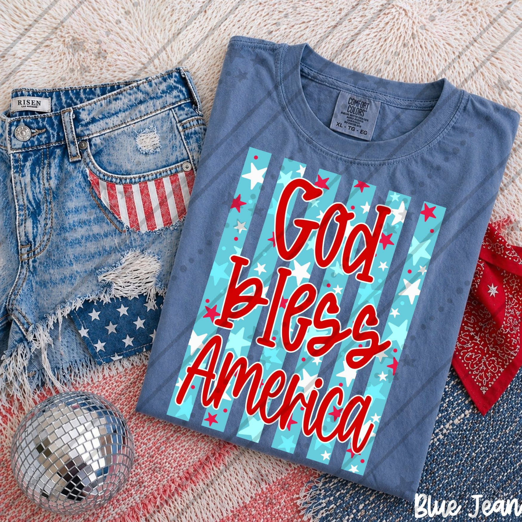 God Bless America Stars & Stripes Comfort Colors patriotic t-shirt featuring red script lettering, turquoise striped American flag-inspired background, and festive Fourth of July Americana style.
