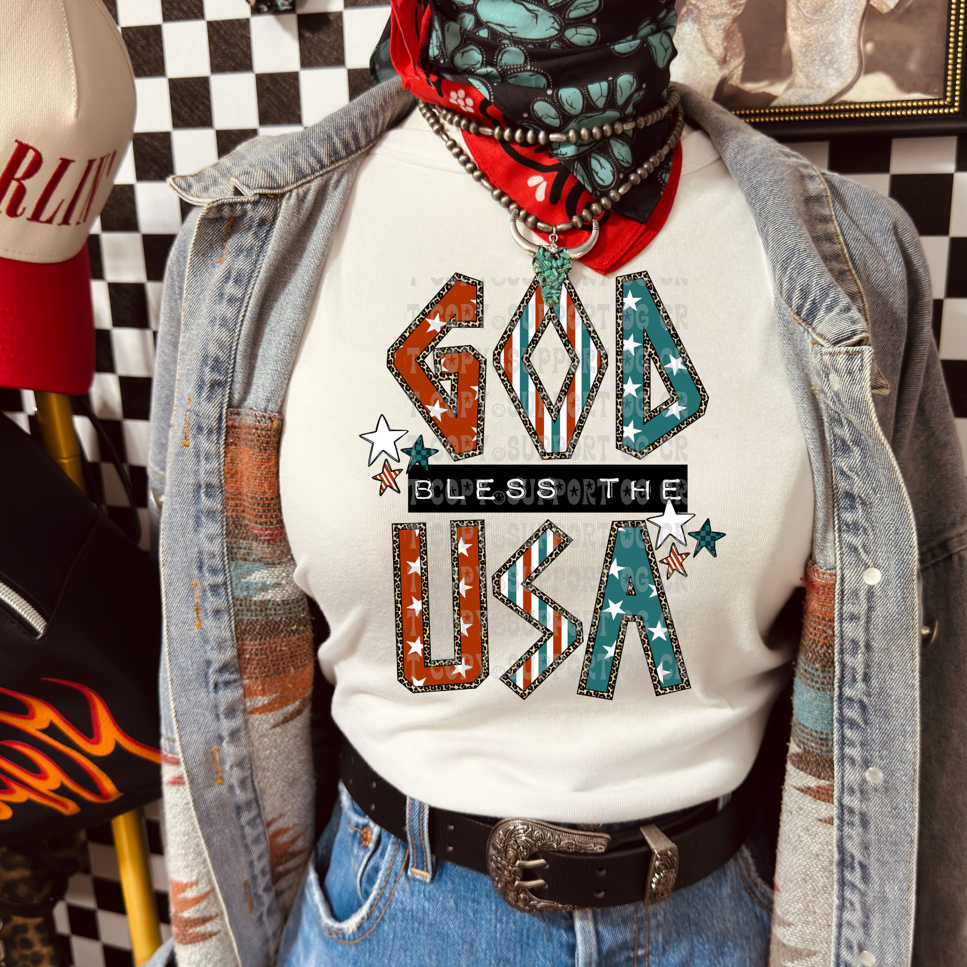 Light heather gray sweatshirt featuring a bold "God Bless the USA" graphic with patriotic stars, vintage red, white, and teal striped lettering, leopard-print accents, and western-inspired details. Styled with denim, an orange wild rag scarf, and western accessories for a trendy Americana look.