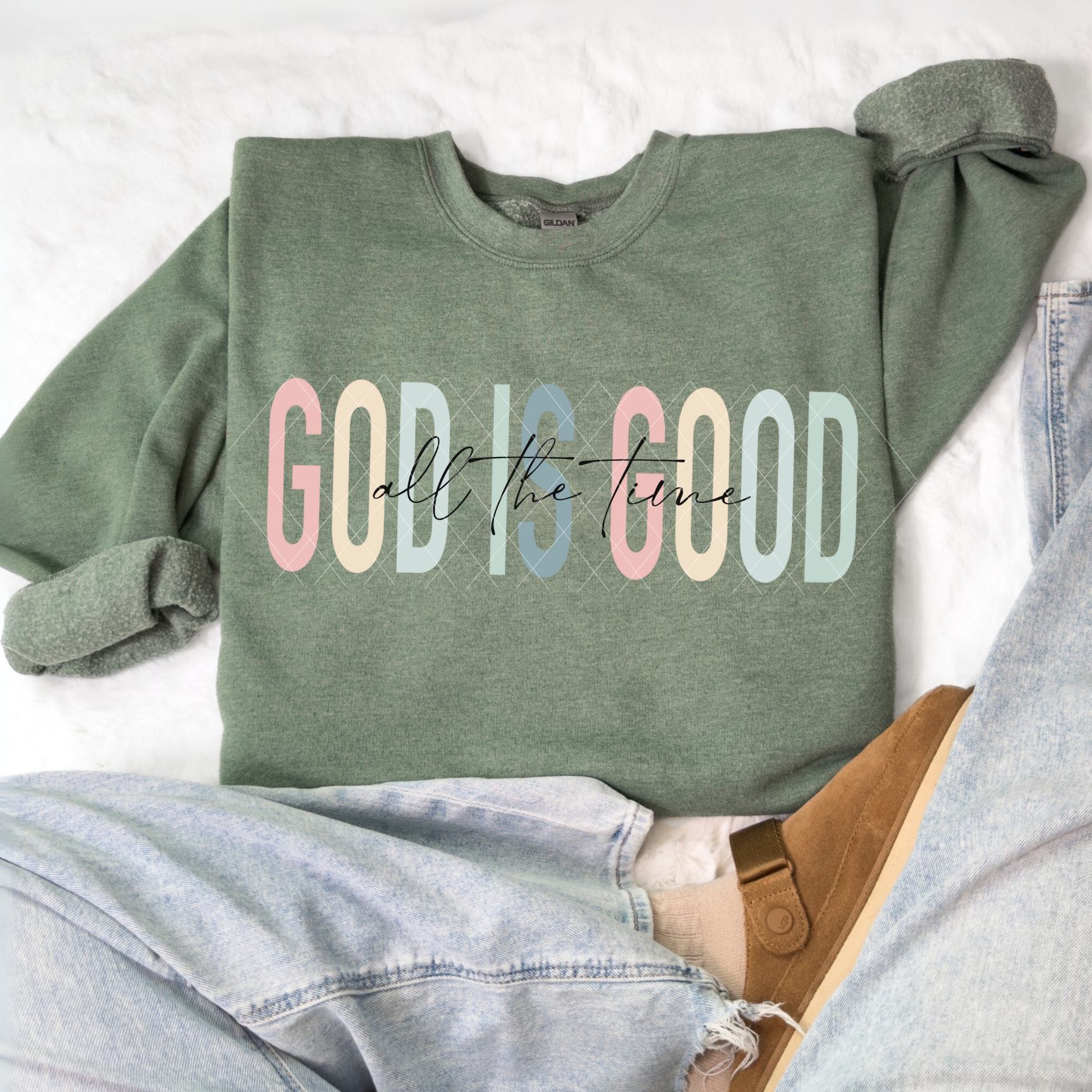 God Is Good All The Time sweatshirt in Gildan Heather Dark Green featuring pastel lettering, flat-lay with denim and slippers.