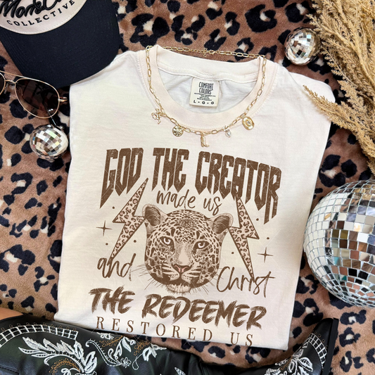 God the Creator Christ the Redeemer graphic tee in Ivory with leopard and lightning bolt design, styled on leopard print backdrop with accessories.