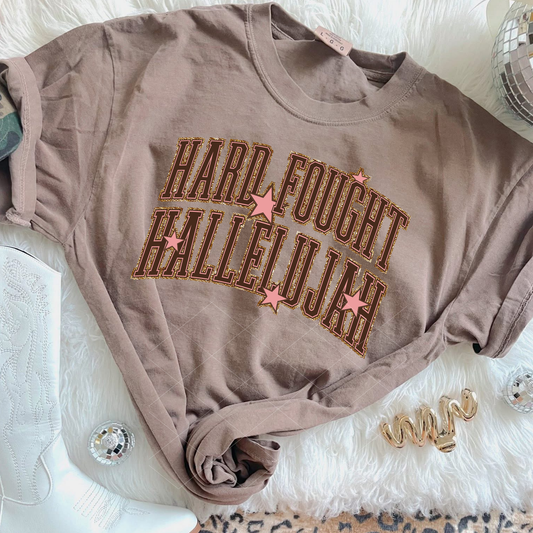 Comfort Colors Espresso Hard Fought Hallelujah tee with distressed collegiate lettering and pink star accents, styled with white Western boots and accessories.