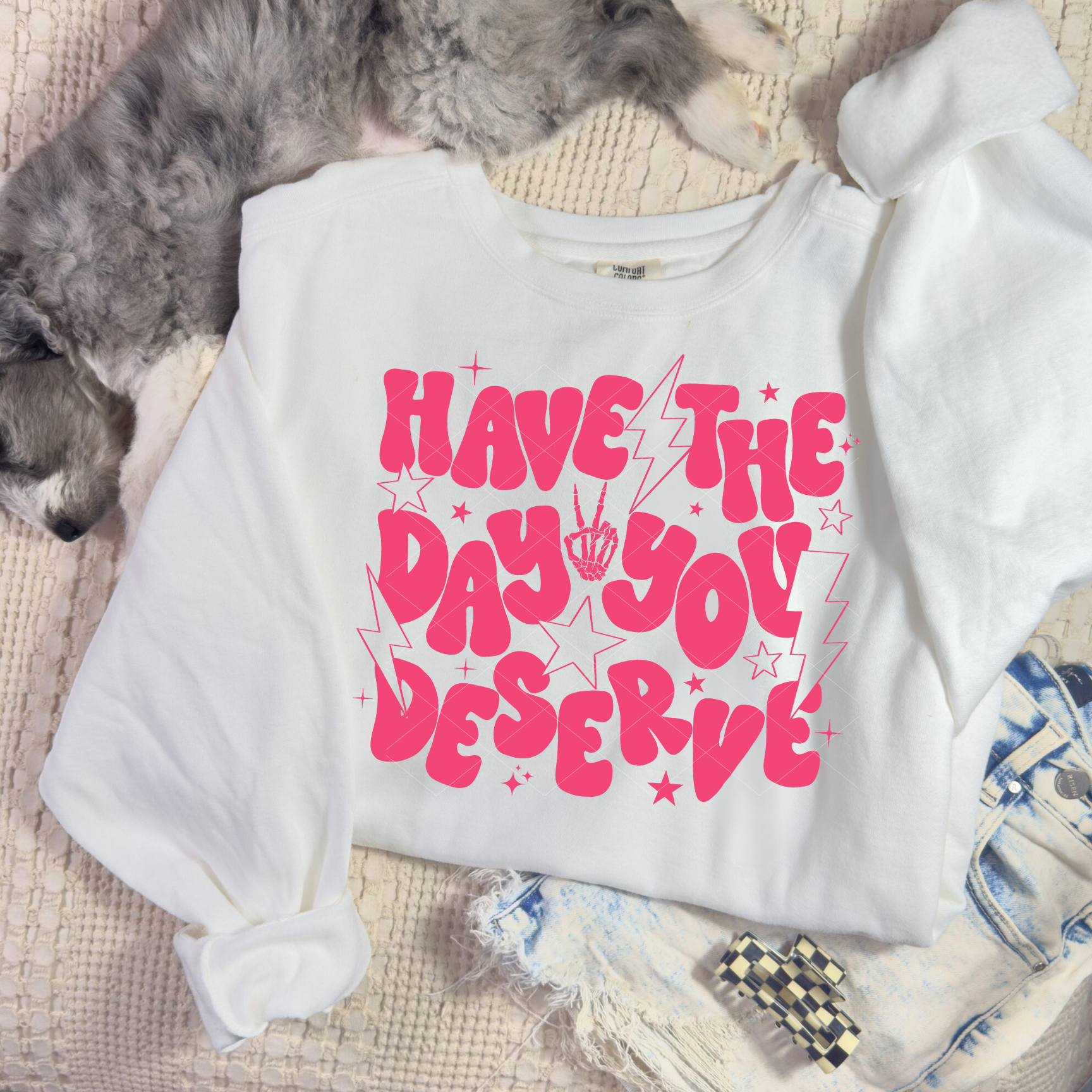 “White ‘Have the Day You Deserve’ sweatshirt with bright pink retro lettering, stars, and lightning bolts, displayed in a flat lay with denim shorts and a dog beside it.