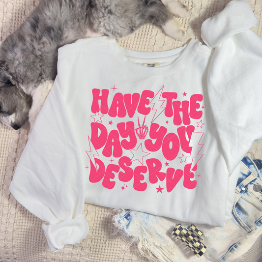 “White ‘Have the Day You Deserve’ sweatshirt with bright pink retro lettering, stars, and lightning bolts, displayed in a flat lay with denim shorts and a dog beside it.