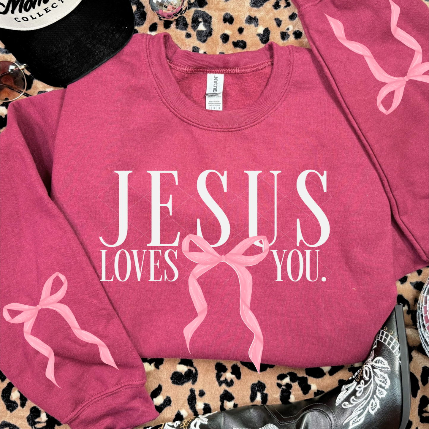 Heather scarlet red Jesus Loves You sweatshirt featuring pink bow design on both sleeves and front graphic, styled with boots and accessories.