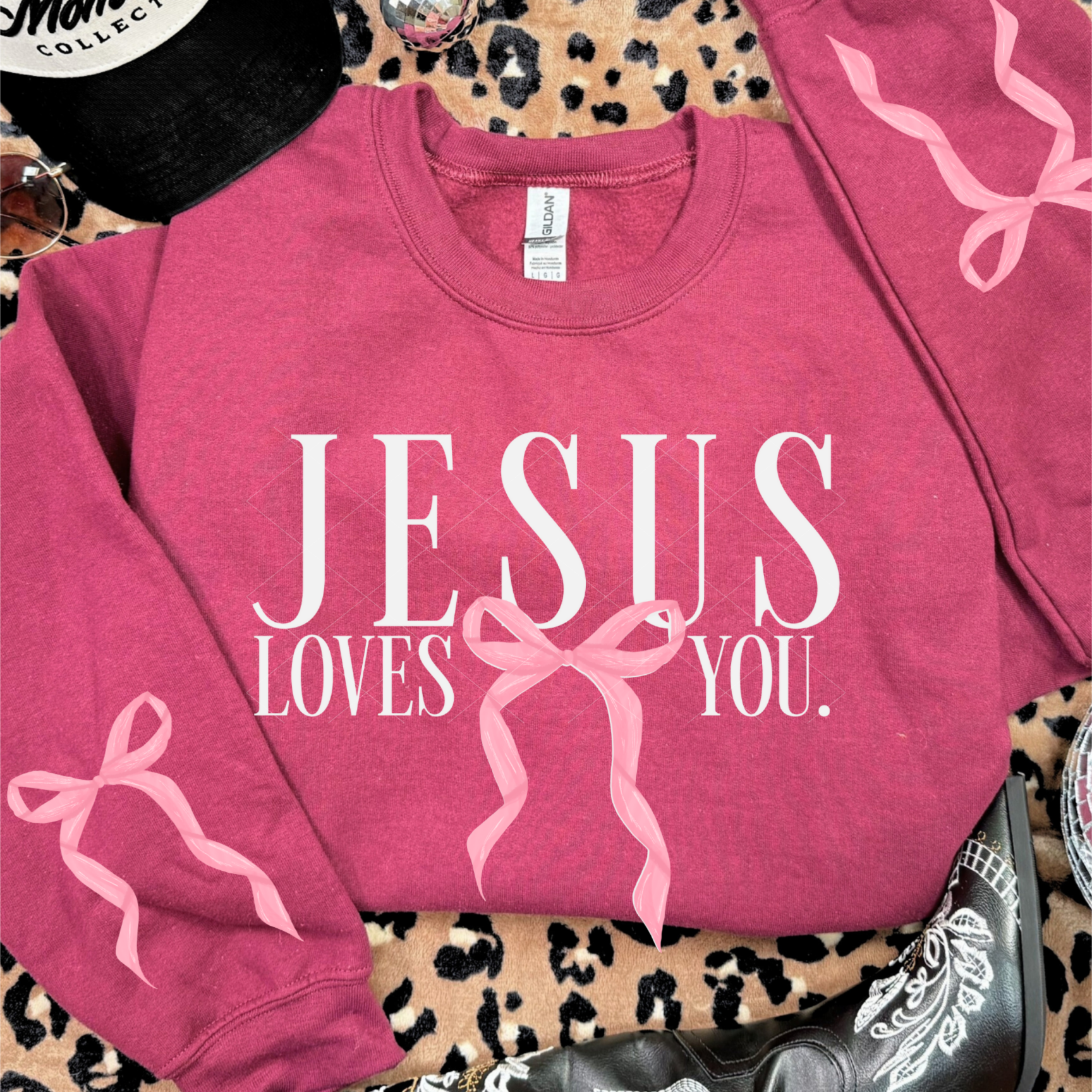 Heather scarlet red Jesus Loves You sweatshirt featuring pink bow design on both sleeves and front graphic, styled with boots and accessories.