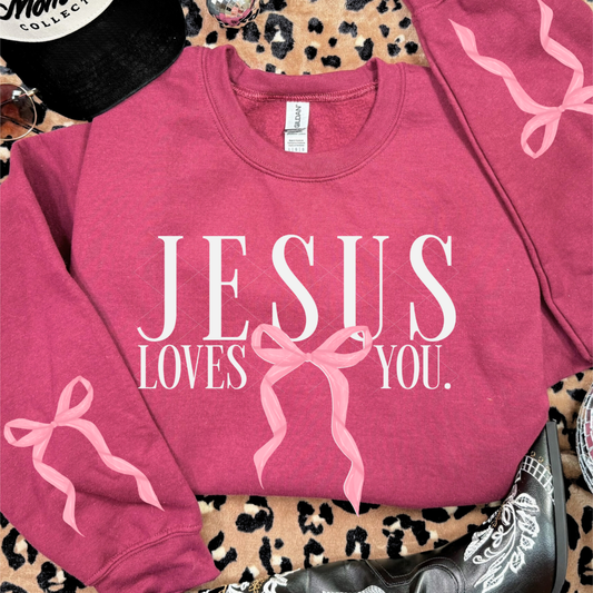 Heather scarlet red Jesus Loves You sweatshirt featuring pink bow design on both sleeves and front graphic, styled with boots and accessories.