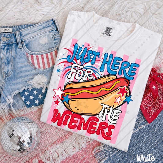 White Comfort Colors® patriotic graphic tee featuring a colorful hot dog illustration with red, white, and blue stars, fireworks, and the phrase "Just Here for the Wiener." Displayed folded with distressed denim shorts featuring an American flag pocket, a red bandana, and a disco ball for a fun Fourth of July-themed flat lay.