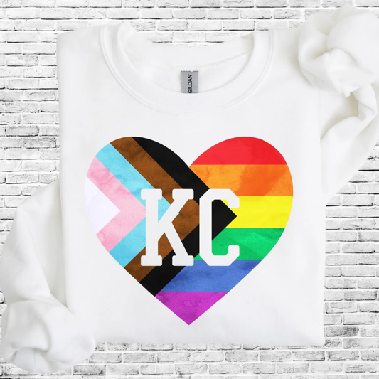 White sweatshirt with KC varsity letters inside an inclusive Pride rainbow heart design, folded against a white brick background.