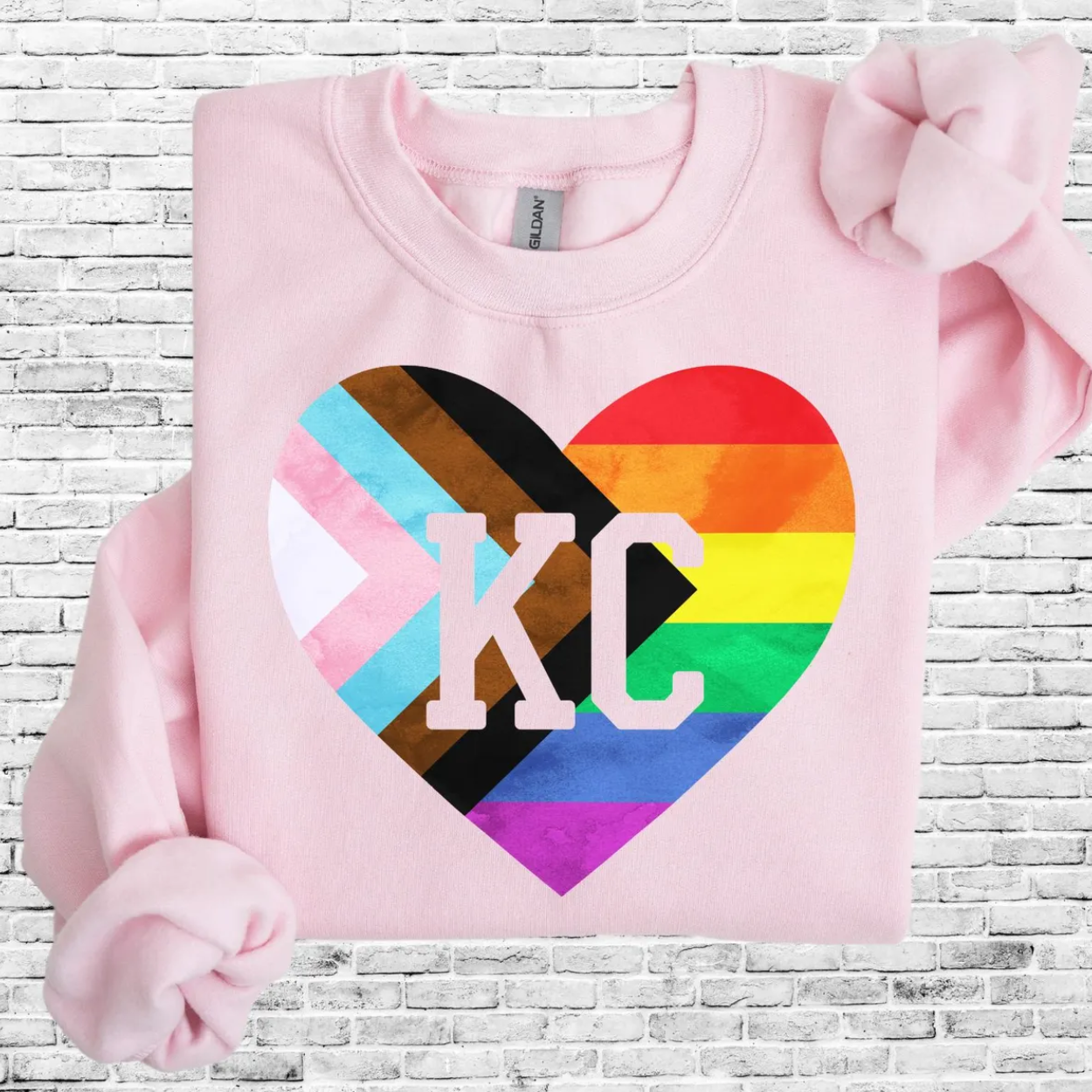 Light pink sweatshirt featuring a KC varsity lettering inside an inclusive Pride rainbow heart graphic, folded on a white brick background.