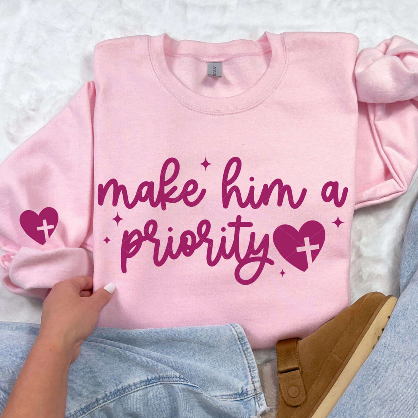 Light pink Make Him a Priority sweatshirt with magenta script and heart-cross design in flat-lay with denim and slippers.