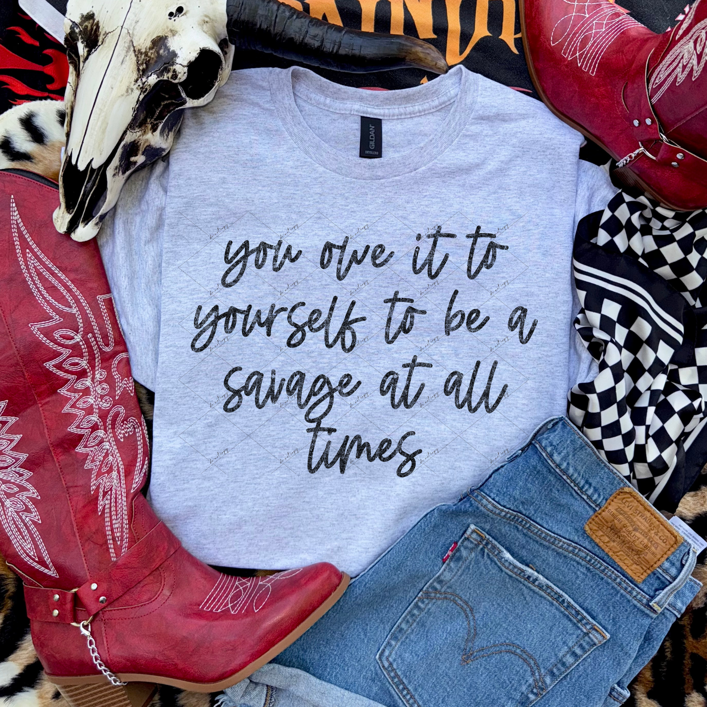Gray graphic tee with the phrase ‘you owe it to yourself to be a savage at all times’ styled with red cowgirl boots, denim jeans, and a decorative skull.