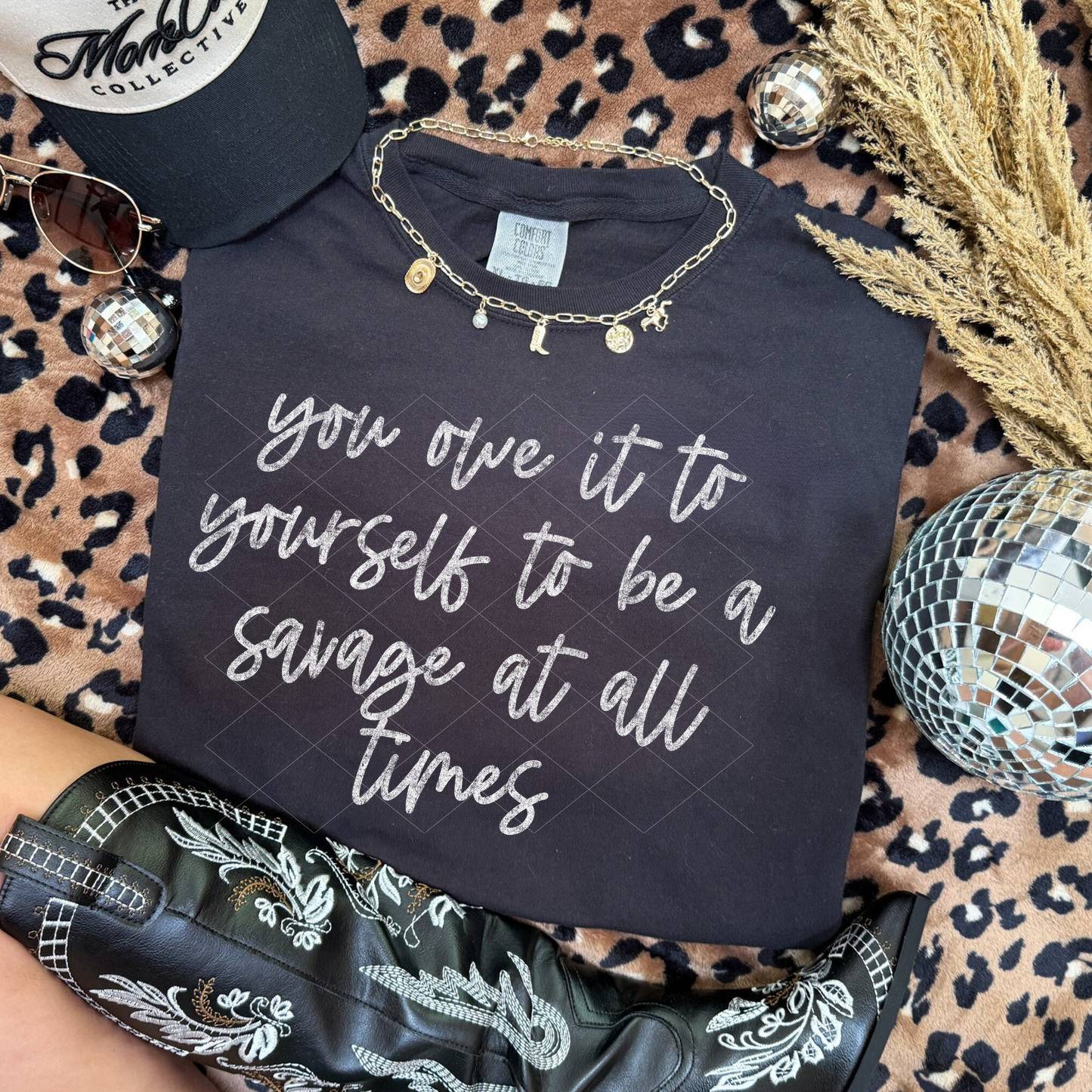 Black graphic tee reading ‘you owe it to yourself to be a savage at all times’ styled with black embroidered boots, a hat, necklace, and disco balls on a leopard background.
