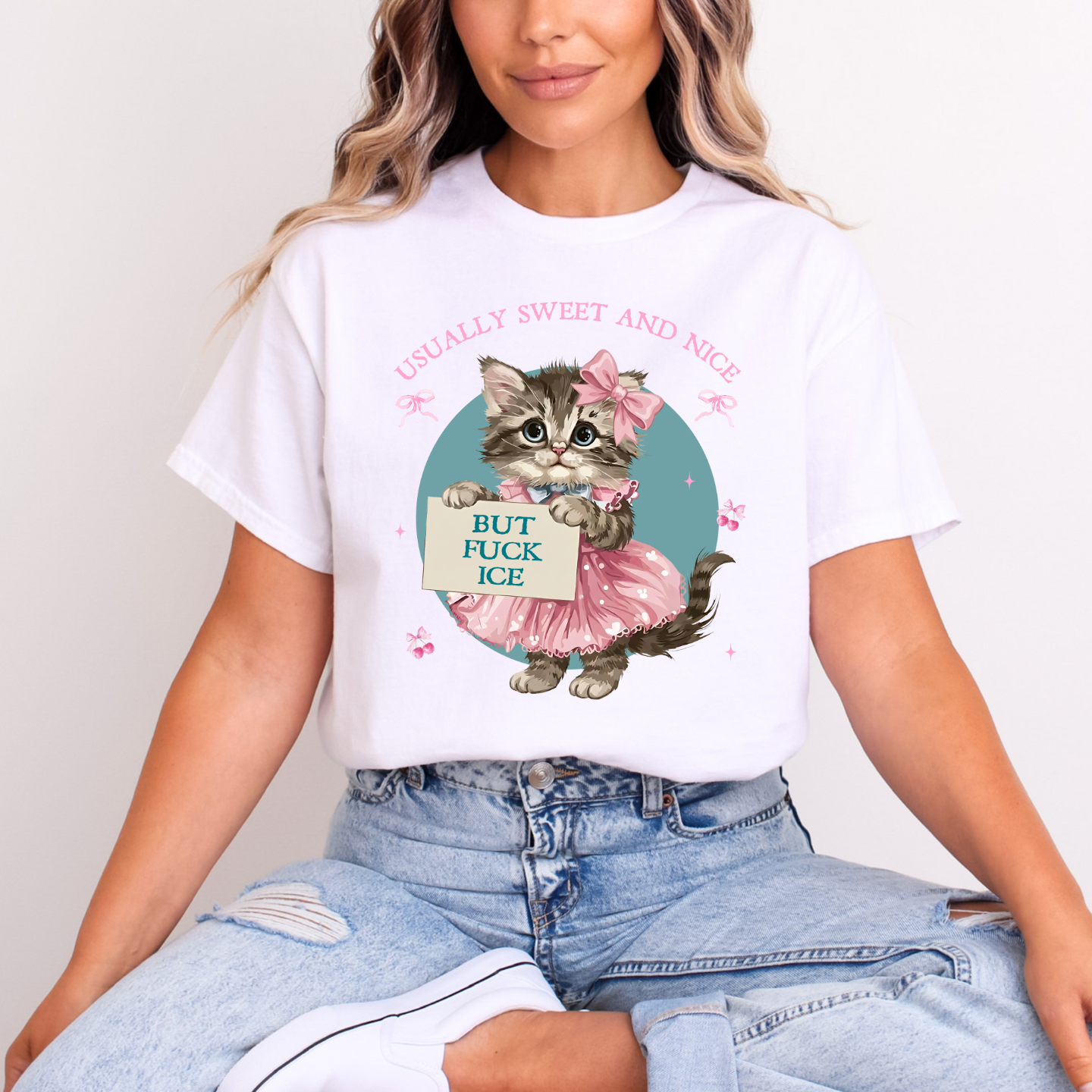 Woman wearing white graphic T-shirt with retro kitten in a pink dress and text ‘Usually Sweet and Nice, But F*ck ICE,’ sitting cross-legged in jeans